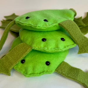 May include: A stack of three plush green bean-shaped toys with black button eyes and felt legs. The toys are made of a textured fabric and are stacked on top of each other.
