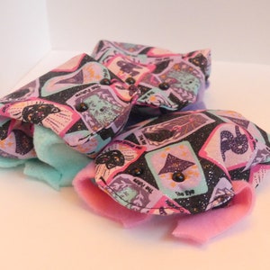 May include: Three fabric pouches with a tarot card design in purple, black, and pink. Each pouch has a light blue interior and a pink flap, secured with a black button. The pouches are arranged on a white surface.