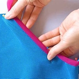 Może przedstawiać: Zbliżenie na rękę przypinającą r&oacute;żową lam&oacute;wkę do niebieskiego materiału. Materiał jest przypinany wzdłuż krawędzi niebieskiego materiału.