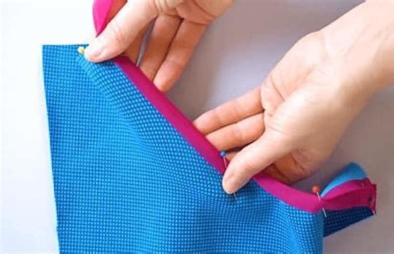 Może przedstawiać: Zbliżenie na rękę przypinającą r&oacute;żową lam&oacute;wkę do niebieskiego materiału. Materiał jest przypinany wzdłuż krawędzi niebieskiego materiału.