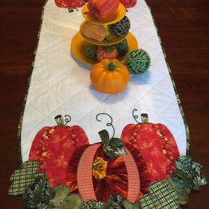 May include: A white quilted table runner with a fall theme. The runner features three pumpkins and green leaves. The pumpkins are made of fabric and have a patchwork design. The leaves are also made of fabric and have a green and brown plaid pattern.