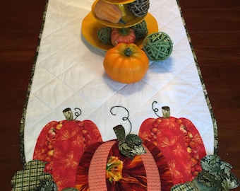 May include: A white quilted table runner with a fall theme. The runner features three pumpkins and green leaves. The pumpkins are made of fabric and have a patchwork design. The leaves are also made of fabric and have a green and brown plaid pattern.