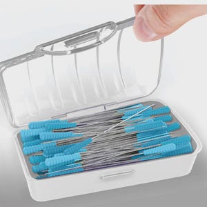 May include: A white plastic case with a clear lid, filled with numerous dental interdental brushes. The brushes have blue, cone-shaped bristles and thin, silver stems. A hand is opening the lid.