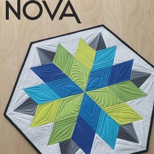 May include: A quilted star-shaped table topper with a black border. The star features shades of blue, green, and gray, with intricate stitching patterns. The word "NOVA" is printed in black at the top. The background is a light wood grain.