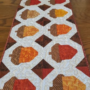 May include: Quilted table runner featuring a repeating pattern of stylized acorns in shades of brown, orange, and red. The acorns are set against a white background with a subtle dotted pattern, and bordered by a brown and red trim. A fall-themed home decor item.