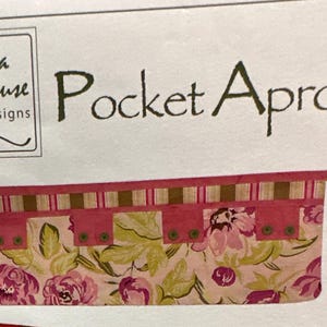 May include: A sewing pattern for a pink and green floral pocket apron with a striped waistband. The pattern is from Vanilla House Designs and is labeled P115 Pocket Aprons.