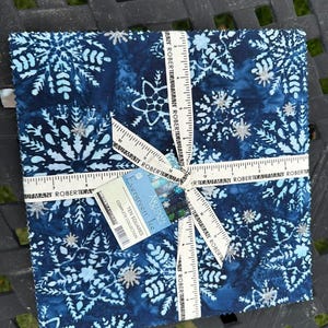 May include: A stack of square fabric pieces featuring a dark blue batik pattern with light blue and silver snowflake designs. A measuring tape marked in inches wraps around the stack, and a tag reads Evergreen Ice Ten Squares Complete Collection.