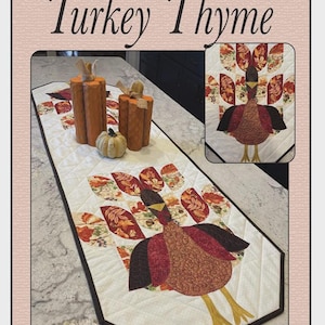 May include: A quilted table runner with a turkey design and the words "Turkey Thyme." The runner features a white background with a diamond pattern and a turkey applique in shades of brown, red, and orange. Decorative pumpkins are also present.