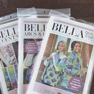 May include: Three sewing pattern packets in clear plastic bags. The packets feature the word "BELLA" in large letters. One packet is labeled "Quilt Coat" and shows two women wearing quilted coats. The other packets are labeled "Accent" and "Arcs & B".