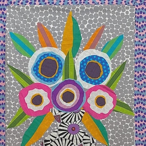 May include: A colorful textile art piece featuring a stylized floral arrangement. The design includes large, circular flowers in shades of pink, purple, and white, with green and yellow leaves. The background is gray with white polka dots, and the border has a dotted pattern.