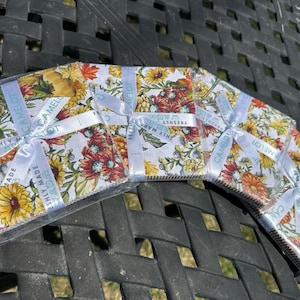 May include: Four stacks of square fabric squares featuring a vibrant floral pattern with yellow, orange, and red flowers on a white background. Each stack is wrapped in clear plastic and tied with a white ribbon printed with the text CAMELOT.