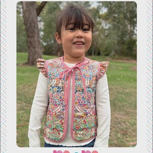 May include: A child wearing a reversible patchwork vest with a floral pattern. The vest has pink trim and a bow closure. The text "HARMONY VEST" and "Melly and Me" are visible.