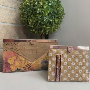 May include: A rectangular cork clutch and a square cork wallet, both featuring rose gold metal frames and clasps. The clutch has a tan cork upper and a floral print lower section. The wallet has a white daisy pattern and a vertical rose gold zipper.