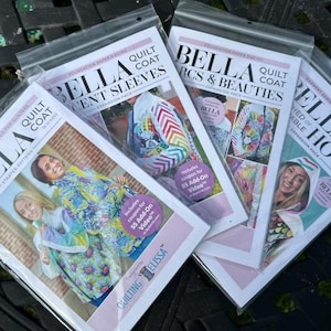 May include: Four Bella Quilt Coat pattern packets in plastic sleeves. The packets feature images of finished coats and the text "Bella Quilt Coat" and "Expansion Pack". The packets are on a dark surface.