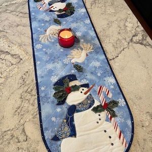 May include: A blue quilted Christmas table runner with a snowman and dove design. The runner features a snowman with a top hat, scarf, and candy cane. Doves and snowflakes are also embroidered on the runner. A red candle sits on the runner.