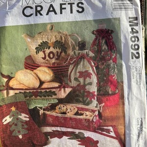 May include: A craft pattern featuring holiday-themed items. Includes a teapot with "Joy" text, bottle bags, oven mitt, and a tray of cookies. The color palette includes red, green, and beige.
