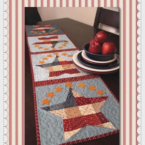 May include: A quilted table runner with a patriotic theme. The runner features a light blue background with star and stripe patterns in red, white, and navy blue. Orange stars are scattered across the surface. The runner is displayed on a dark wooden table.