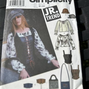 May include: A Simplicity sewing pattern envelope, number 5799, featuring illustrations of a long-sleeved blouse, a corset-style vest, a hat, and a bag. The cover displays a model wearing the blouse and vest, with text including Simplicity, JR. TREND, and size AA 3/4-9/10.