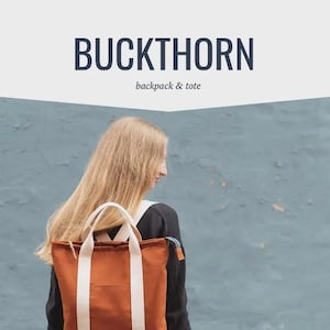 May include: A rust-colored backpack and tote bag with cream-colored straps. The bag has a front pocket and a zipper closure. The word "BUCKTHORN" is printed in dark blue above the words "backpack & tote". The background is a muted blue.