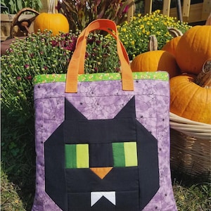 May include: A Halloween-themed tote bag featuring a black cat face design. The bag has a purple background with a spiderweb pattern, green and orange accents, and orange handles. Pumpkins and flowers are in the background.
