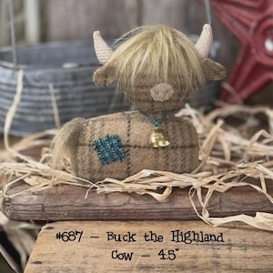 May include: A plush Highland cow figurine made of tan plaid fabric with a fuzzy tan mane, two small horns, and a teal patch on its side. A small gold bell hangs from a teal cord around its neck. Text on the base reads #687 - Buck the Highland Cow - 4.5".