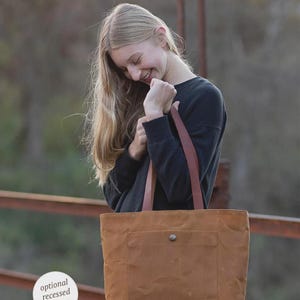 May include: A brown tote bag with a front pocket and brown leather straps. The bag has a button closure and the text "optional recessed zipper". The bag is held by a person.