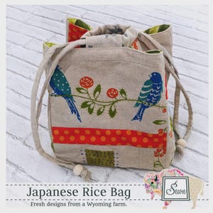 May include: A beige fabric Japanese rice bag featuring a print of two blue birds perched on a branch with red flowers. The bag includes a horizontal orange band with white polka dots, a green patterned square patch, and two drawstring handles with wooden beads.