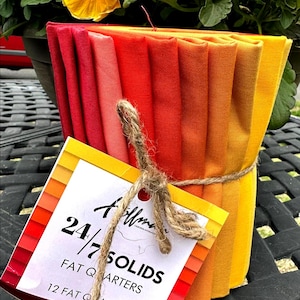 May include: A bundle of 12 fat quarters in a gradient of red, orange, and yellow hues, tied with twine. A tag with the text "24/7 Solids" and "Fat Quarters" is attached. Yellow flowers are in the background.