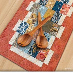 May include: A colorful patchwork table runner with a geometric design in shades of blue, orange, yellow, and green. The runner is made of fabric squares sewn together. Two wooden spoons are placed on top of the runner.