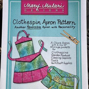 May include: A sewing pattern for a Clothespin Apron by Mary Mulari Designs. The pattern features a pink and green apron design with pockets, and the text "Clothespin Apron Pattern" is displayed. The apron is described as reversible and suitable for various supplies.