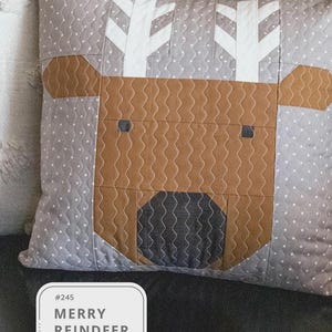 May include: A square decorative pillow with a reindeer design. The reindeer face is brown with a black nose and gray eyes, set against a gray background with white polka dots. White tree-like shapes are at the top. The text "MERRY REINDEER" is in a white box.