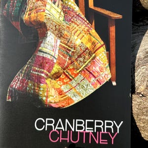 Puede incluir: Una colorida colcha de retazos, drapeada sobre una silla de madera. La colcha presenta una variedad de patrones y colores, incluyendo rojo, naranja, amarillo y verde. El texto "CRANBERRY CHUTNEY" se muestra en blanco y rosa.