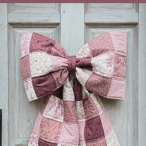 May include: A large, quilted bow in shades of pink and burgundy hangs on a light gray door. The bow is made of patchwork squares with floral and solid patterns. The door has a simple, paneled design and a dark handle.