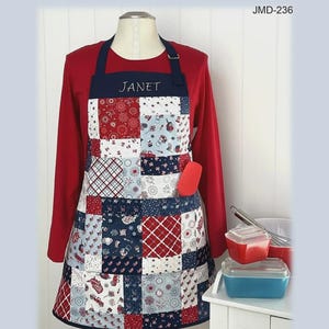 May include: A patchwork apron featuring a red, white, and blue color scheme with various patterns including stars, stripes, and small motifs. The apron has a dark blue neck strap, a front pocket, and the name JANET embroidered on the chest. Size 26 inches wide by 29 inches high.