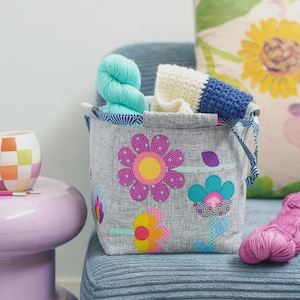May include: A gray fabric storage basket featuring colorful floral applique designs in pink, yellow, and blue. The basket is filled with a skein of bright turquoise yarn and a folded piece of cream and navy blue crocheted fabric.