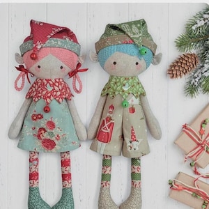 May include: Two handmade elf dolls with fabric hats, dresses, and legs. One doll has a pink hat and dress with red floral accents, the other has a green hat and dress with red and white accents. The dolls are on a white wooden surface.