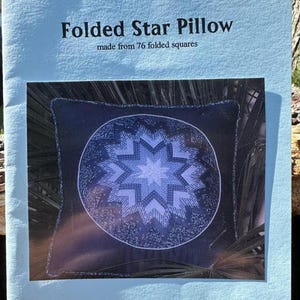 May include: A light blue instructional booklet titled Folded Star Pillow made from 76 folded squares. The cover features a photograph of a square dark blue pillow with a central circular design composed of layered blue and white star patterns.