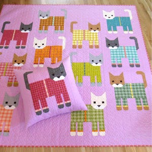 May include: A purple quilt with a pattern of cats wearing pajamas. The cats are in various colors and patterns, including red, blue, green, yellow, and orange. There is a pillow in the center of the quilt with a cat wearing red pajamas.