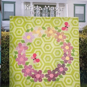 May include: A quilt pattern titled "Flutterby Frolic" by Krista Moser. The quilt features a floral design with pink and purple flowers and butterflies on a green hexagonal background. Sizes include twin (76" x 84"), wall (48" x 56"), and table runner (12.5" x 41").