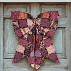 May include: A large, handmade rag quilt bow in shades of burgundy, mauve, and tan, with a patchwork design. The bow is made of various fabric squares and hangs against a weathered, light blue door. The text reads "THE BIG GIANT RAG QUILT BOW".