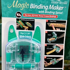 May include: A clear and green Magic Binding Maker with Binding Spool by Taylor Seville. The package includes instructions and images demonstrating the EZ-Fold and Sharp-Crease Binding process. It is designed for 2 1/4" wide fabric.