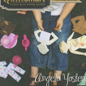 May include: A Quiltsmart printed interfacing product package titled Fusible Flats by Angela Yosten. The cover features a person holding a fabric doll with blue jeans and a white shirt, surrounded by toy tea sets and additional fabric doll pieces.