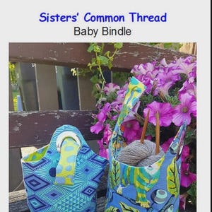 May include: Two fabric baby bindles with blue and green patterns. One has a handle with a yellow and green polka dot design. The other holds gray yarn and knitting needles. The text "Sisters' Common Thread Baby Bindle" is at the top.
