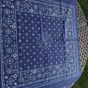 May include: A square navy blue bandana featuring a white and light blue floral and geometric paisley-inspired pattern. The design includes a central diamond motif surrounded by smaller floral elements and a decorative border along the edges.