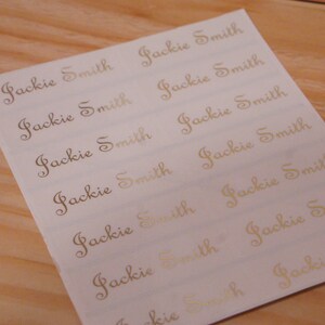 May include: A sheet of white labels with the name "Jackie Smith" printed in a gold, elegant script. The labels are arranged in rows and columns, and the sheet is placed on a light brown wooden surface.