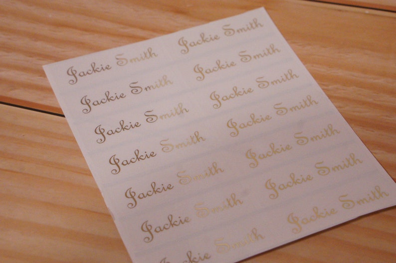 May include: A sheet of white labels with the name "Jackie Smith" printed in a gold, elegant script. The labels are arranged in rows and columns, and the sheet is placed on a light brown wooden surface.