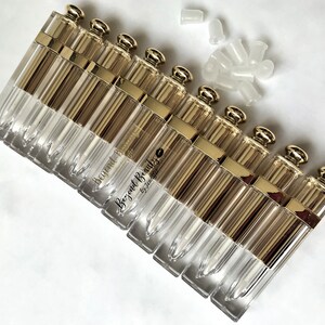 May include: A collection of clear, rectangular lip gloss tubes with gold-colored caps and accents. The tubes are arranged in a row, with several white plastic applicators scattered nearby. The text "Beyond Beauty by Jennifer" is printed on some of the tubes.