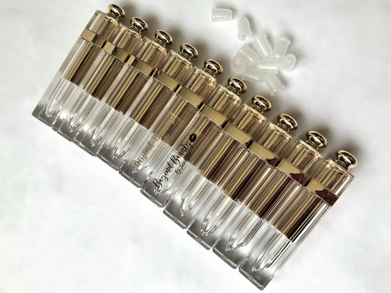 May include: A collection of clear, rectangular lip gloss tubes with gold-colored caps and accents. The tubes are arranged in a row, with several white plastic applicators scattered nearby. The text "Beyond Beauty by Jennifer" is printed on some of the tubes.