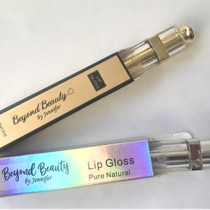 May include: Two lip gloss tubes in rectangular boxes. The boxes are black and gold with the words "Beyond Beauty by Jennifer" and "Lip Gloss" printed on them. The lip gloss tubes are clear with gold accents and a gold cap. The boxes read "Net: 5ml".
