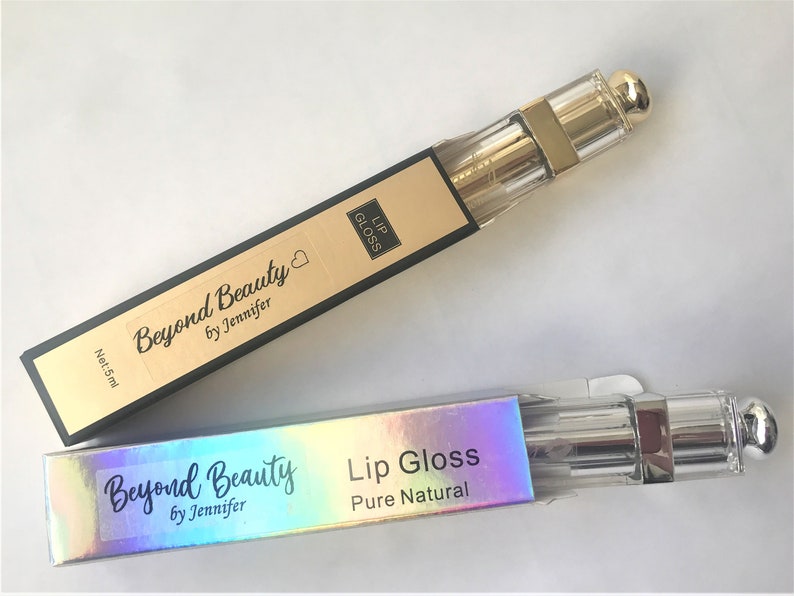 May include: Two lip gloss tubes in rectangular boxes. The boxes are black and gold with the words "Beyond Beauty by Jennifer" and "Lip Gloss" printed on them. The lip gloss tubes are clear with gold accents and a gold cap. The boxes read "Net: 5ml".
